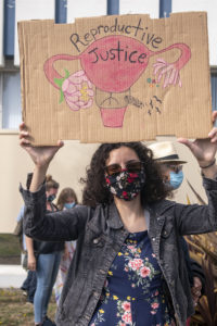 Graciela Leal holds a cardboard sign with 'Racial Justice' written on a uterus.