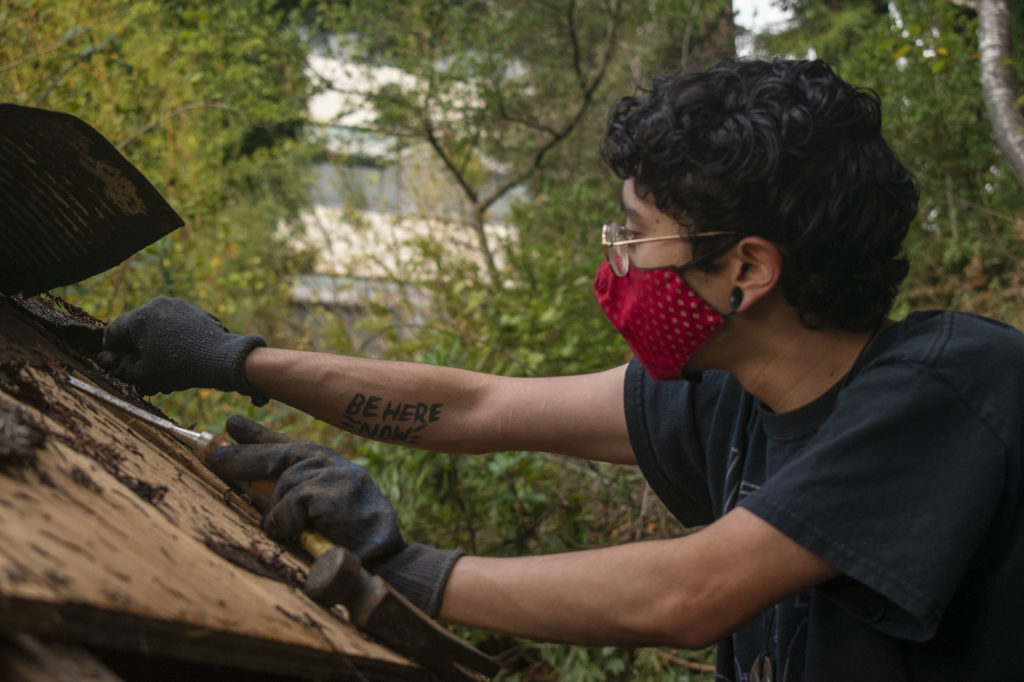 James Lara removes shingles on the yurt roof. Text on his arm reads 'Be Here Now'.