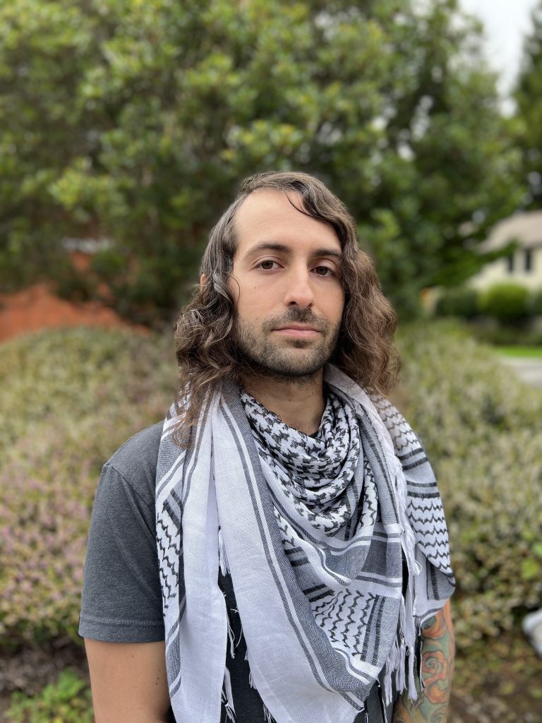Rick Toledo wearing kuffia in front of Siemens Hall. Green bushes behind him. 