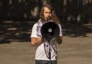 Rick Tolelendo holding megaphone. Wearing white shirt and has long curly brown hair.
