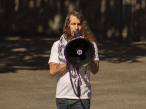 Rick Tolelendo holding megaphone. Wearing white shirt and has long curly brown hair.