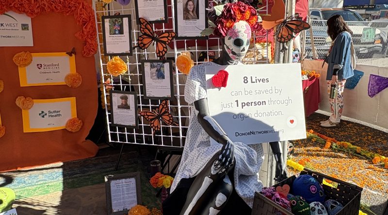 An altar is shown at a Dia de muertos festival. There is a skeleton in front of the altar holding a sign that says, "8 lives can be saved by just every 1 person through organ donation."